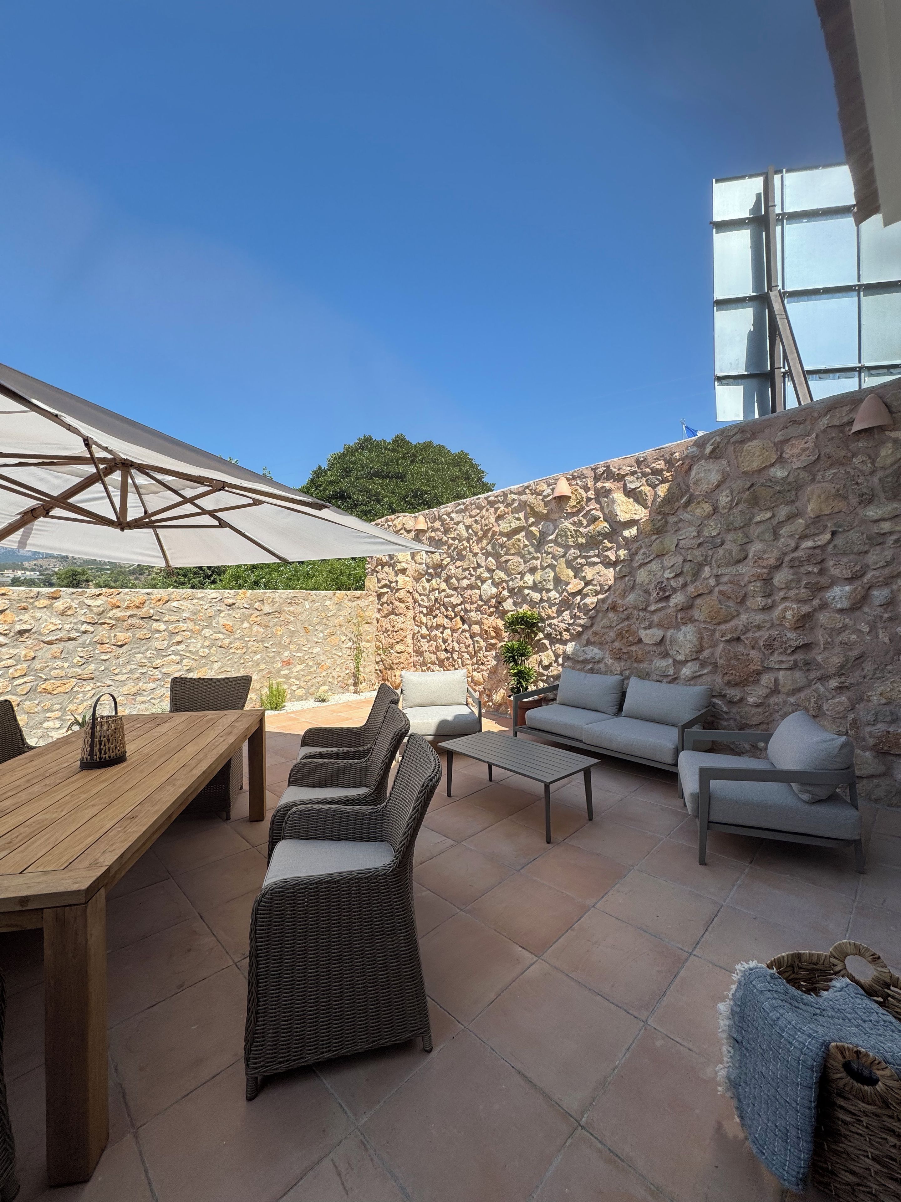 Un patio al aire libre con pared de piedra, mobiliario de mimbre, una mesa de madera y un paraguas grande.