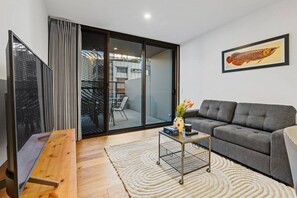 Un salon moderne avec un canapé gris, une table basse en bois ornée d’un vase de fleurs, une porte-fenêtre coulissante donnant sur un balcon et un tableau représentant des poissons, encadré, accroché au mur.