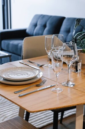 Une table à manger dressée avec des assiettes, des couverts et des verres à vin, avec un canapé et une plante en arrière-plan.