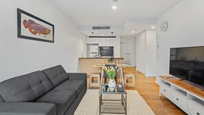 Una sala de estar moderna con un sofá gris, una mesa de centro con un florero y un televisor, con una cocina al fondo.