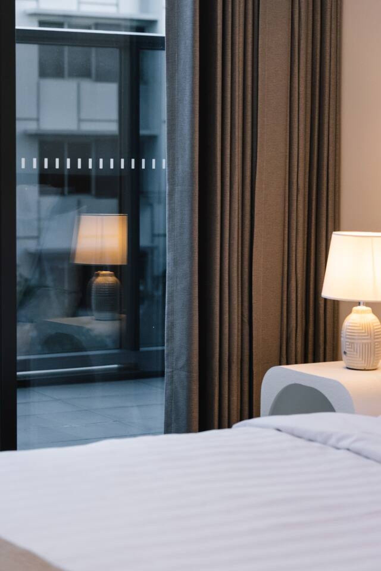 Een hotelkamer met een bed, een nachtkastje met een lamp en uitzicht op een gebouw door een raam met gordijnen.