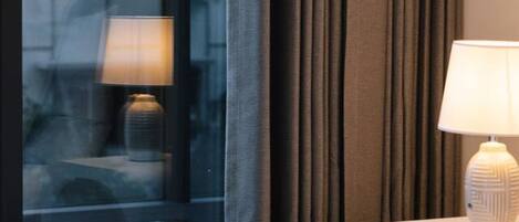 Habitación de hotel con una cama, una mesita de noche con lámpara y vistas a un edificio a través de una ventana con cortinas.