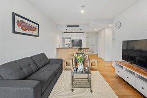 Ein modernes Wohnzimmer mit einem grauen Sofa, einem Couchtisch mit Blumen in einer Vase, einem Fernseher auf einem Holzboden und einer Küche im Hintergrund.