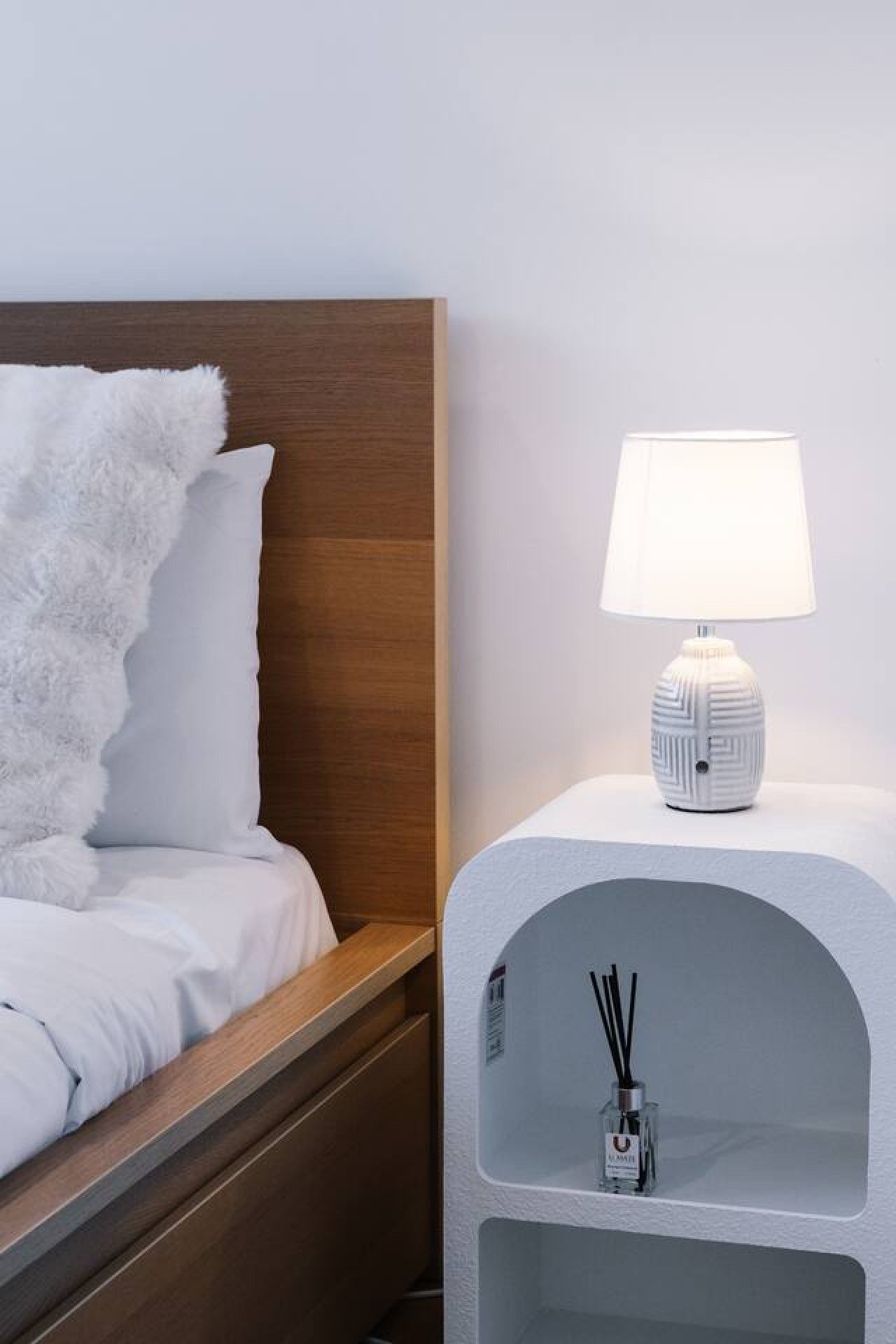 Un lit bien fait, avec une literie blanche et un plaid blanc moelleux, une tête de lit en bois et une table de chevet sur laquelle se trouve une lampe blanche et une petite étagère avec un flacon de diffuseur à bâtonnets.