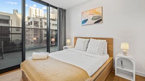 Habitación de hotel moderna con una cama grande, dos mesitas de noche con lámparas, vistas a la ciudad a través de ventanales de gran altura y un cuadro en la pared.