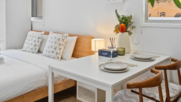 Une chambre d’hôtel avec un lit, une table blanche ornée d’un vase, d’une bougie et d’une boîte de mouchoirs.