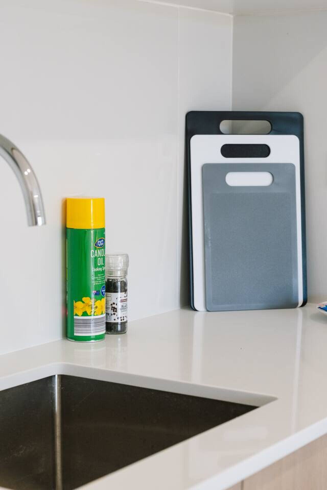 Una cocina con encimera blanca, un lavamanos de acero inoxidable, un recipiente amarillo y verde de aceite en aerosol, una botella pequeña de sal y una tabla de cortar gris.