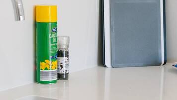 A kitchen with a white countertop, a stainless steel sink, a yellow and green canister of cooking spray, a small bottle of salt, and a gray cutting board.