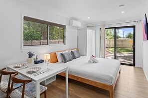 Ein modernes Schlafzimmer mit Bett, Esstisch mit Stühlen, einem Fenster und einer Glastür zu einem Außenbereich.