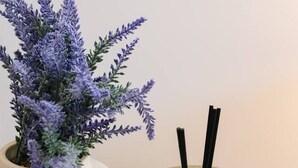 Eine klare Glasflasche mit Etikett, ein Kerzenhalter aus Glas und eine Vase mit lila Blumen.