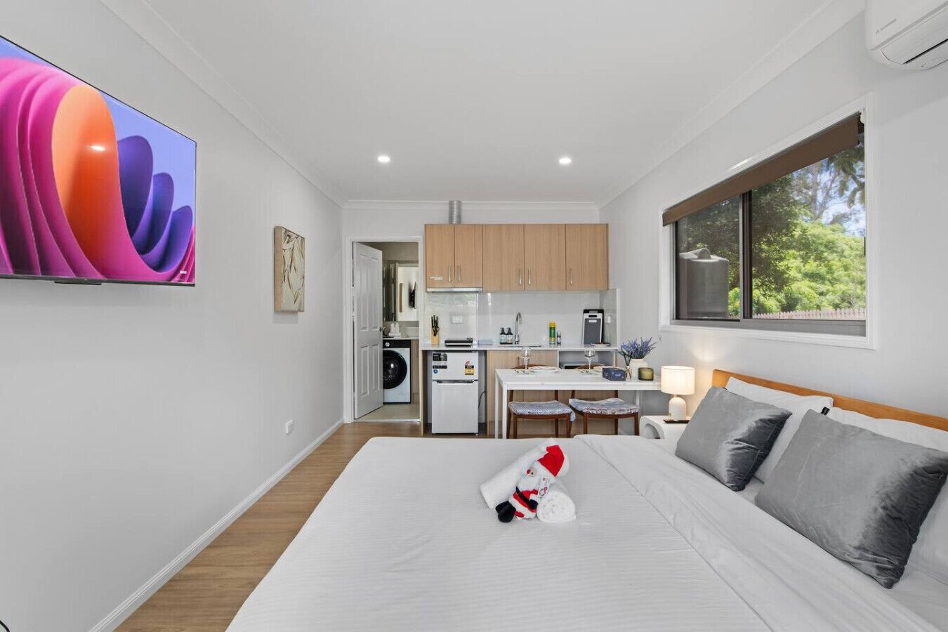A modern hotel room with a large bed, a small kitchenette, a dining area, and a view of greenery outside.