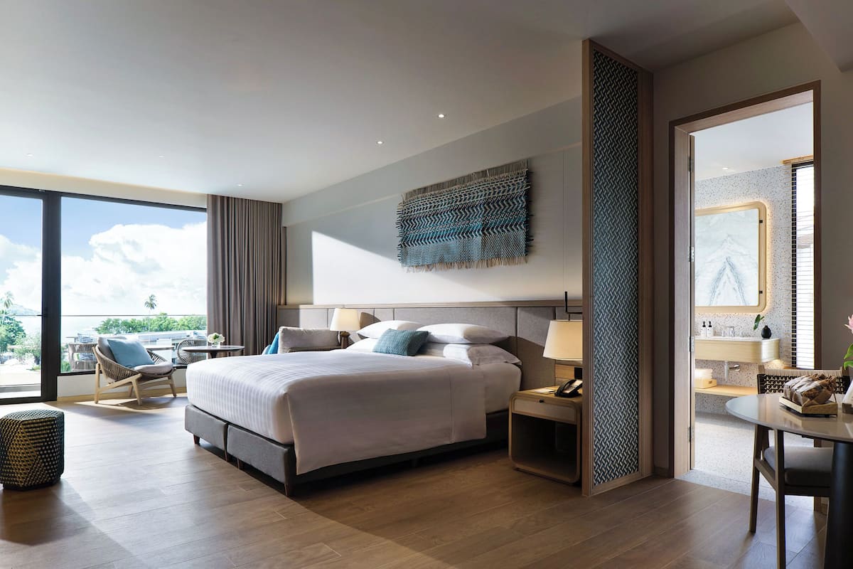 A modern hotel room with a large bed, bedside tables, a chair, a desk, and a view of the outdoors.