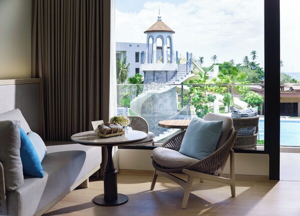 Habitación de hotel moderna con balcón, una mesa de comedor y vistas a un complejo turístico con fuente de agua y palmeras.