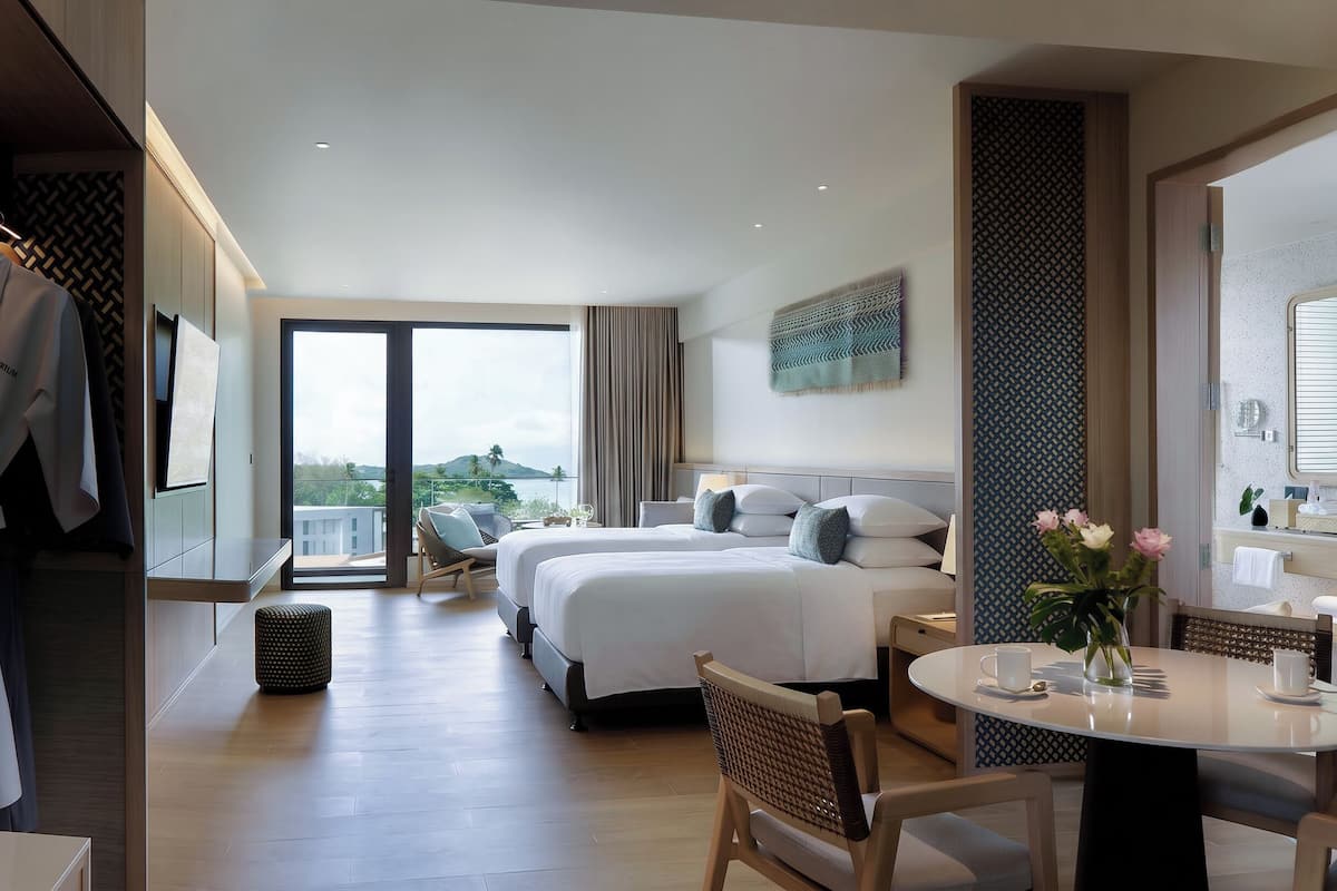 A hotel room with a large bed, a dining table, a chair, a small stool, a wardrobe, and a view of the outdoors.