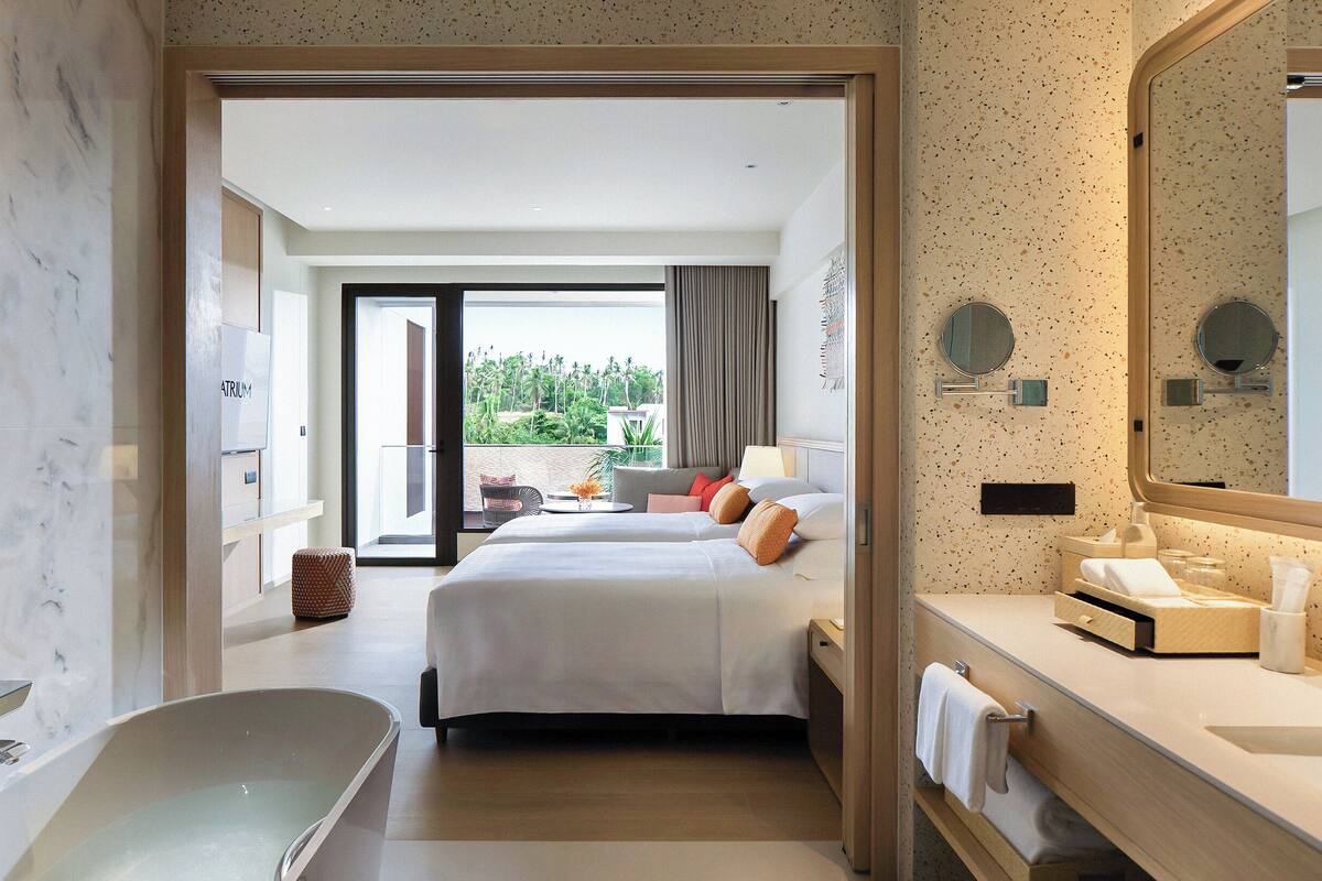 A hotel room with a large bed, a bathtub, a bathroom with a sink, and a view of palm trees.