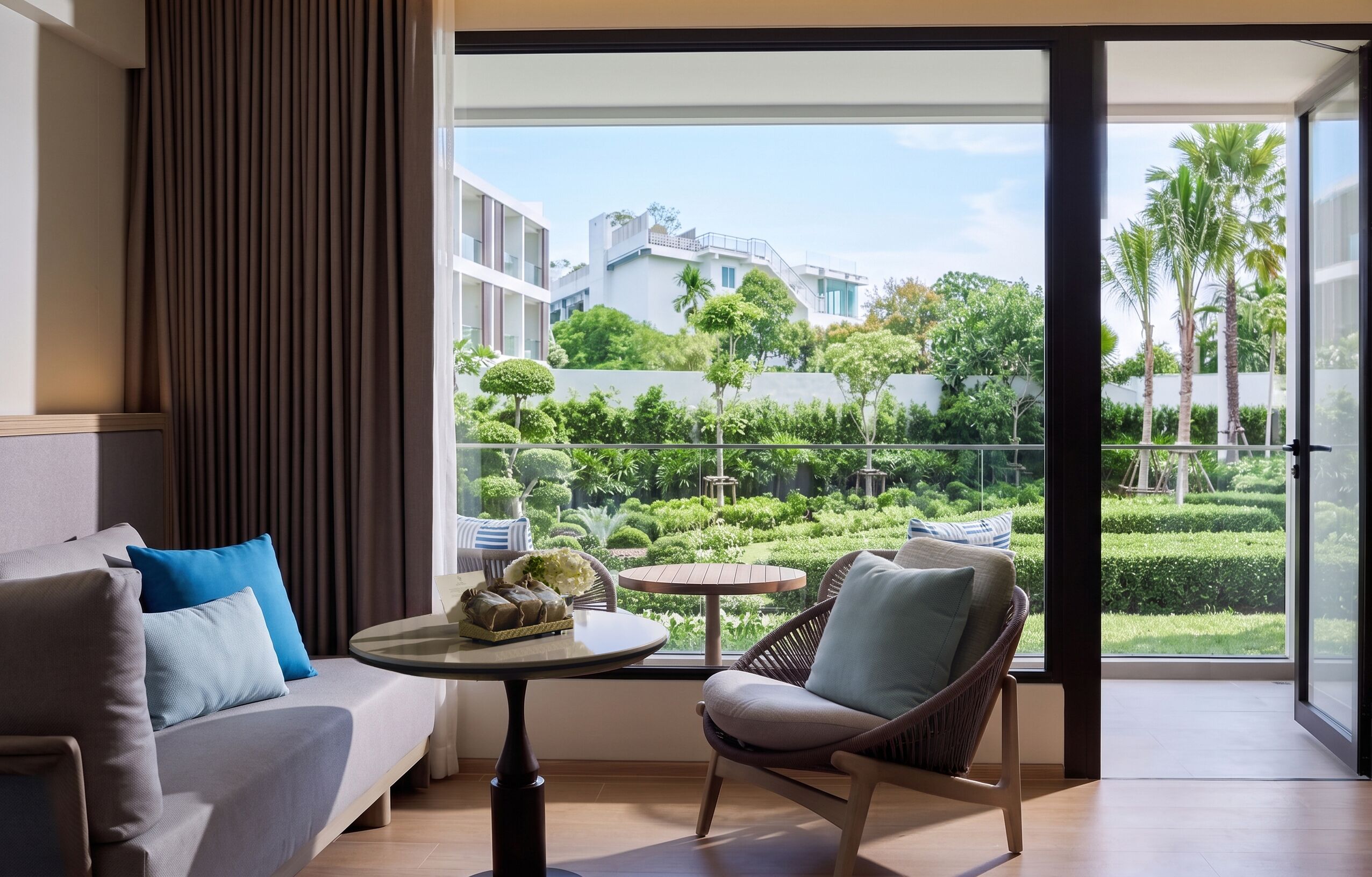 A modern living room with a sofa, chair, and table, a balcony with a view of greenery and buildings.