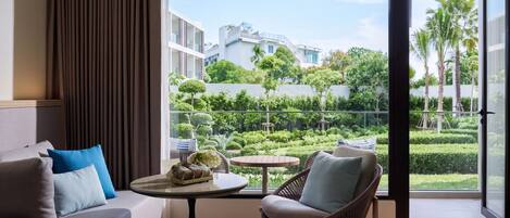 A modern living room with a sofa, chair, and table, a balcony with a view of greenery and buildings.