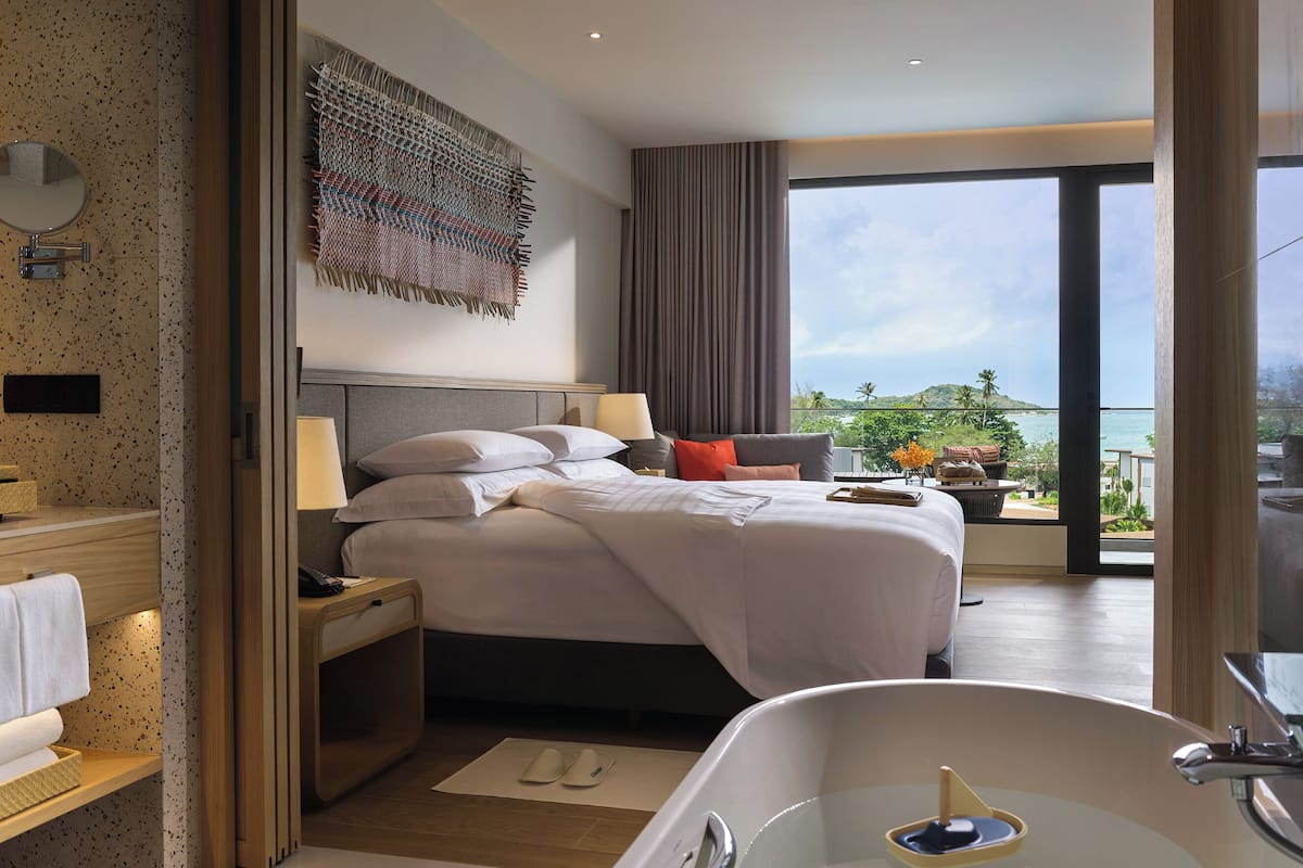 A modern hotel room with a large bed, a bathtub, a balcony with a view, and a wall-mounted mirror.