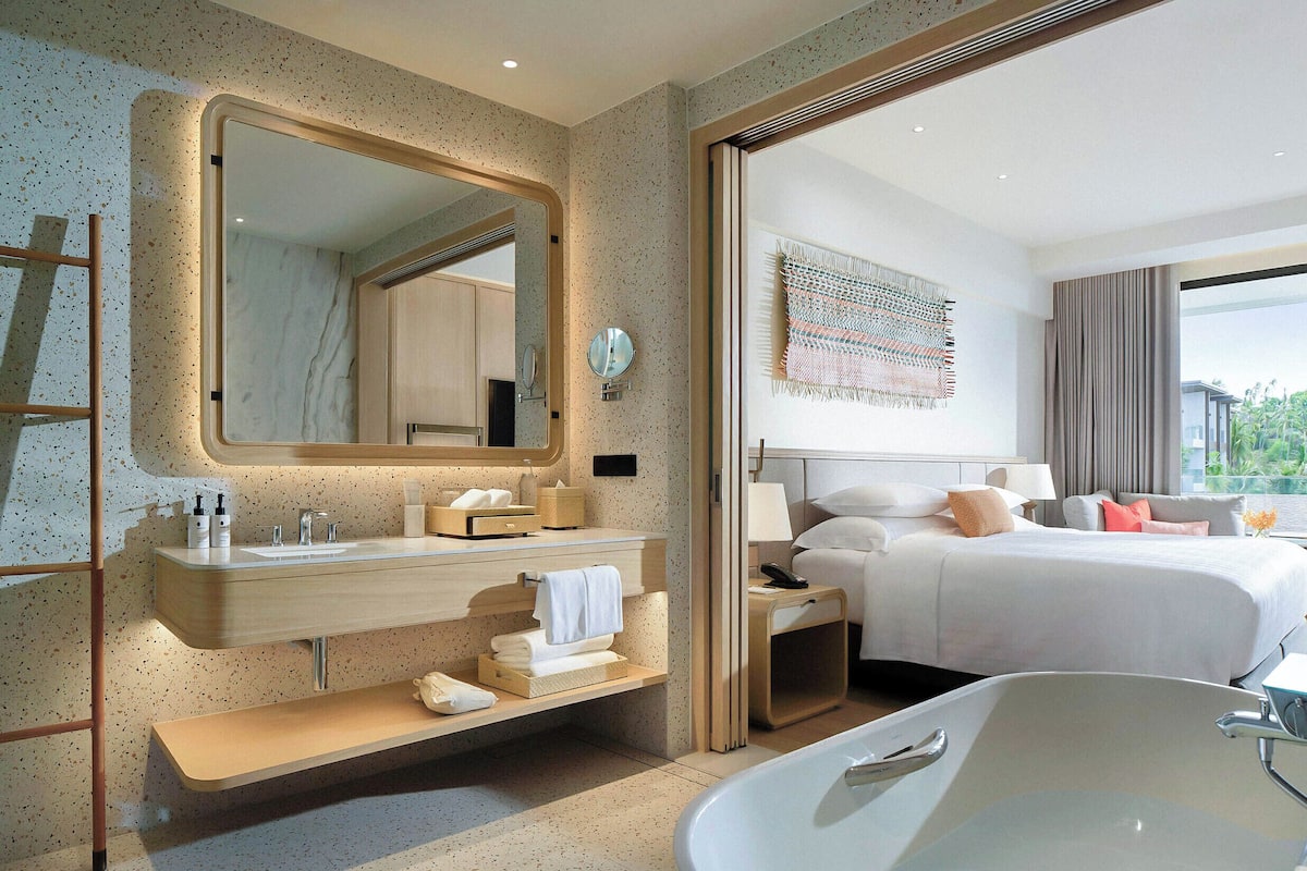 A modern hotel room with a large bathtub, a wooden vanity with a mirror, a bed with pillows, and a view of the outdoors.
