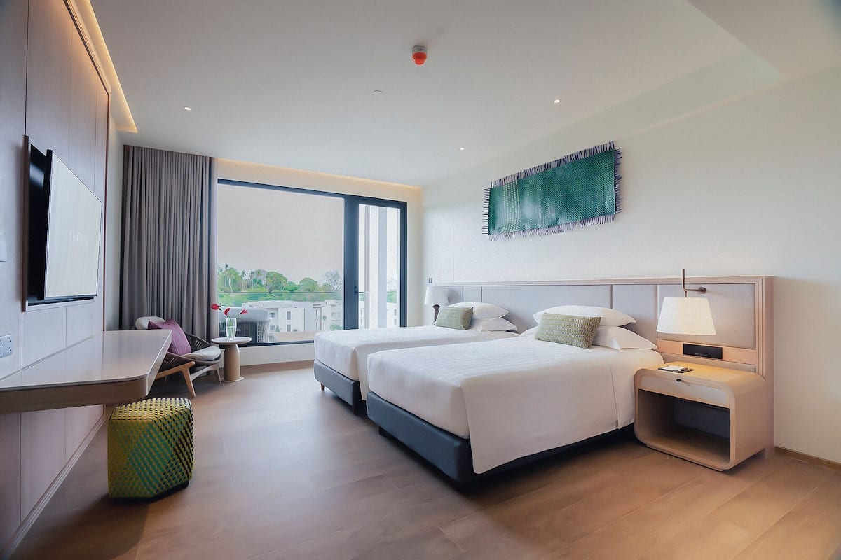 A modern hotel room with a large bed, bedside tables, a desk, and a view of greenery outside.