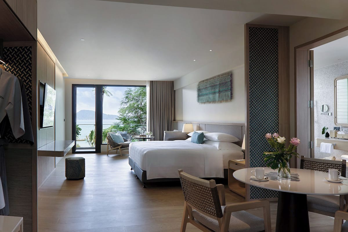 A hotel room with a bed, bedside tables, a chair, a round table with a vase of flowers, and a view of the outdoors.