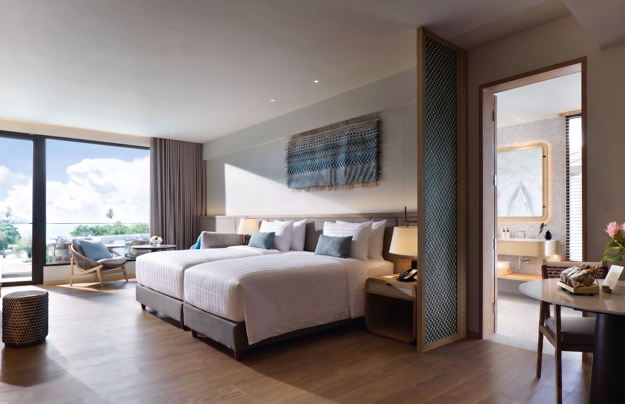 A modern hotel room with a large bed, a sitting area with a chair, a small table, and a view of the outdoors.
