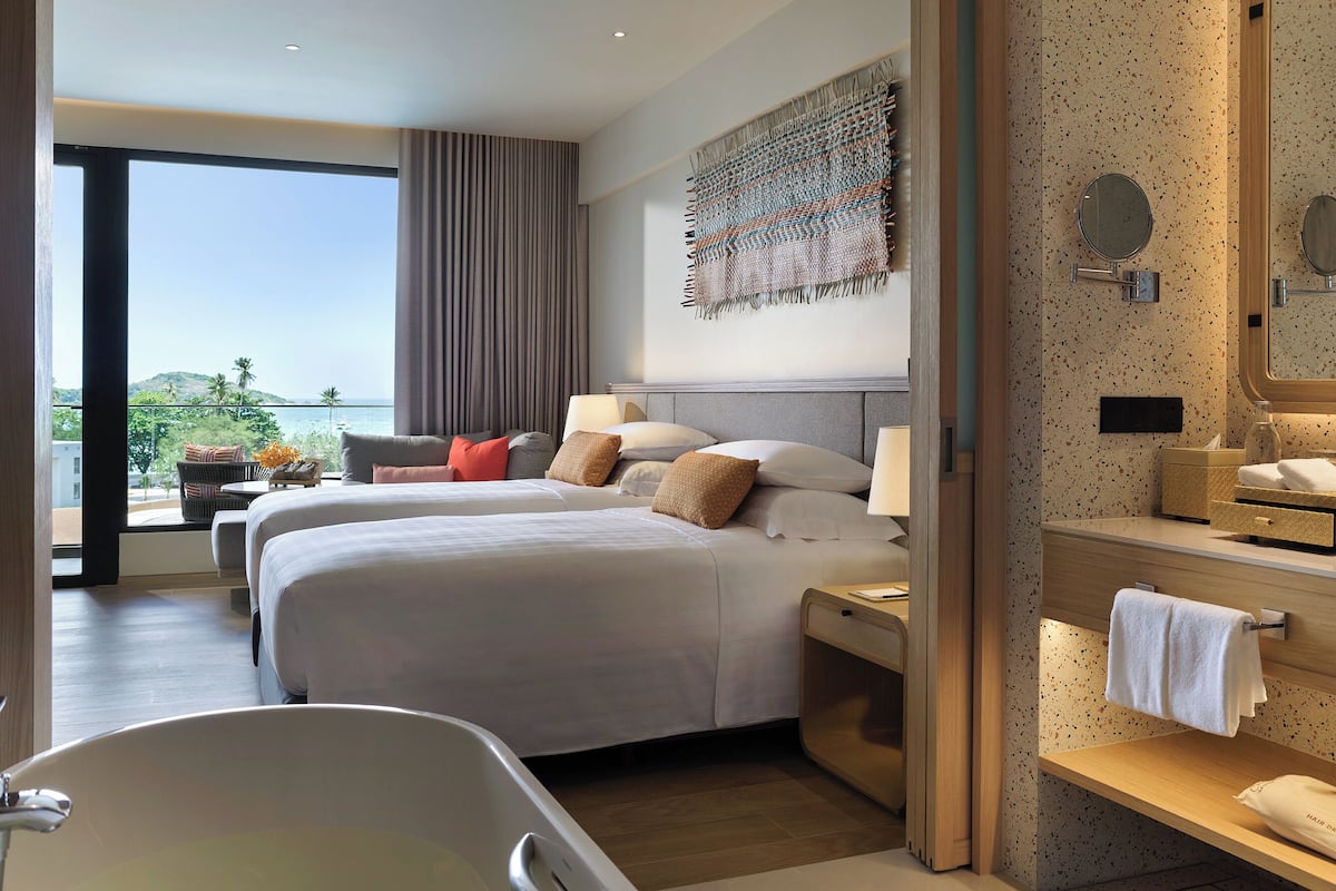 A hotel room with a large bed, a bathtub, a bathroom with a mirror, and a view of the sea.
