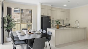 A modern kitchen with a central island, a dining area with a black table and grey chairs, and a view of the outside through a large window. - Spacious 4BR Family Home in Inala | Quiet Street (Inala)