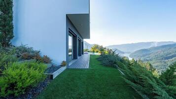 Una casa moderna con un camino de entrada largo y estrecho que conduce a una terraza de madera, rodeada de exuberante vegetación y con vistas a un paisaje montañoso.
