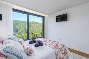 A bedroom with a large window, a bed with floral bedding, and a TV mounted on the wall. - Gerês Paradise (Vieira do Minho)