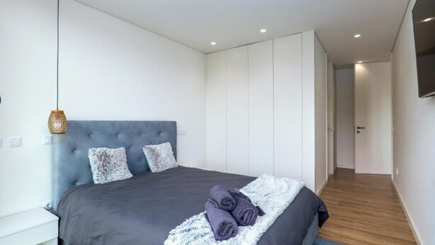Quarto moderno com uma cama grande, cabeceira estofada cinza e uma TV de tela plana na parede.