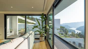 A modern balcony with a sofa, a potted plant, and a view of a lake and mountains.