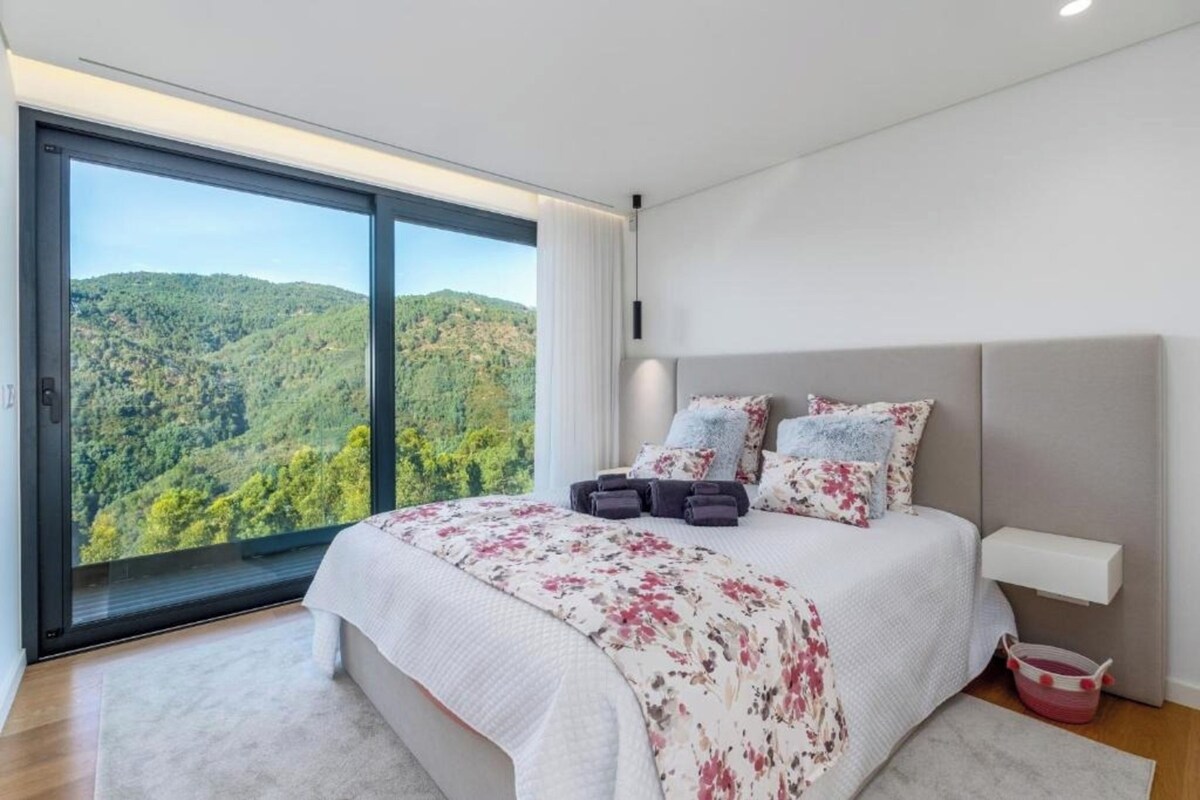 A bedroom with a large bed, a nightstand, a bedside table, and a view of a forested landscape through a sliding door.