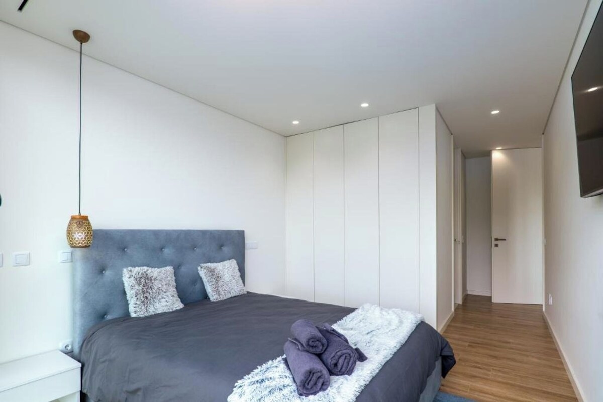 A modern bedroom with a large bed, a grey upholstered headboard, and a flat-screen TV mounted on the wall.