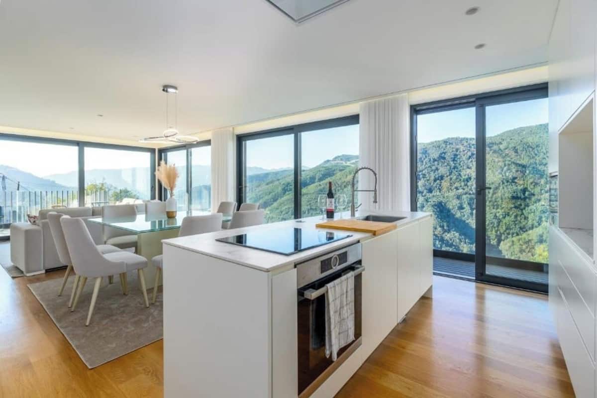 A modern kitchen with a central island, large windows, and a dining area with white furniture.