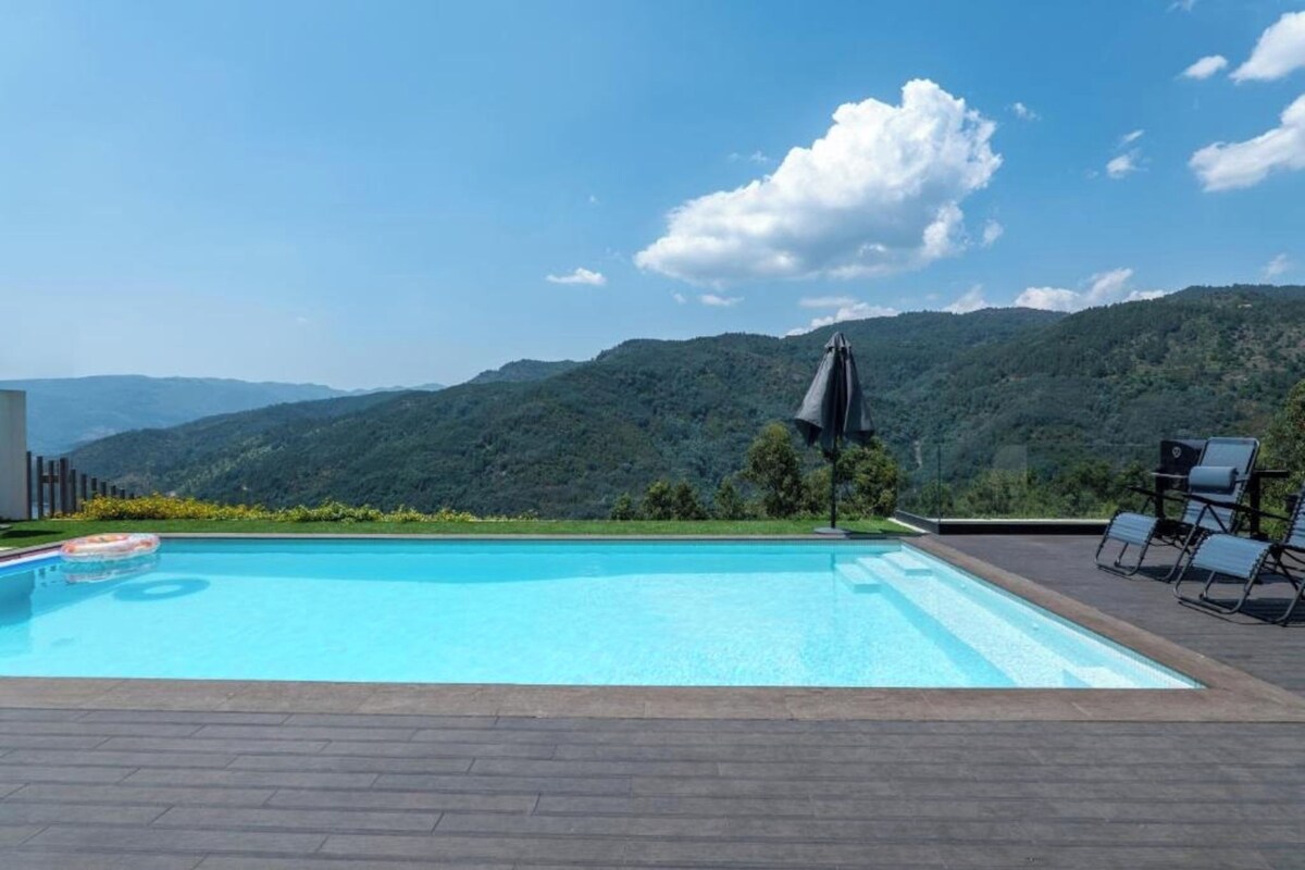 A swimming pool with a view of mountains, surrounded by lounge chairs and an umbrella.