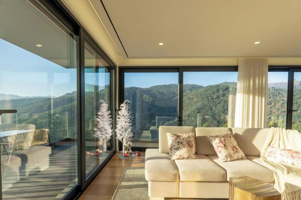 A modern living room with a large glass wall, a beige sofa, and a view of a mountainous landscape.