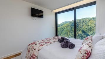 A bedroom with a large window, a bed with a floral bedspread, and a rolled towel on the bed.