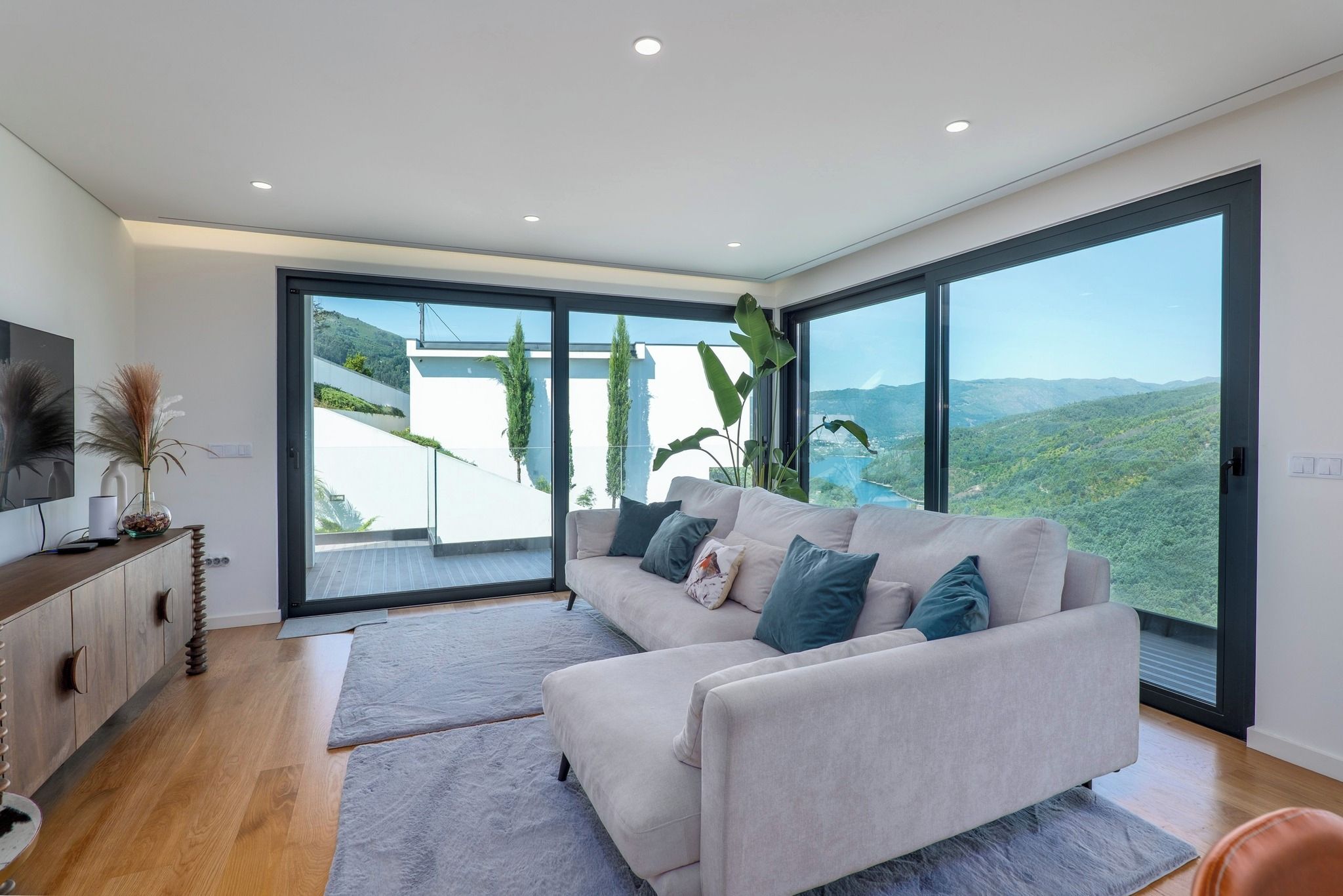 A modern living room with a large sofa, a flat-screen TV, and a view of a mountainous landscape through large windows.