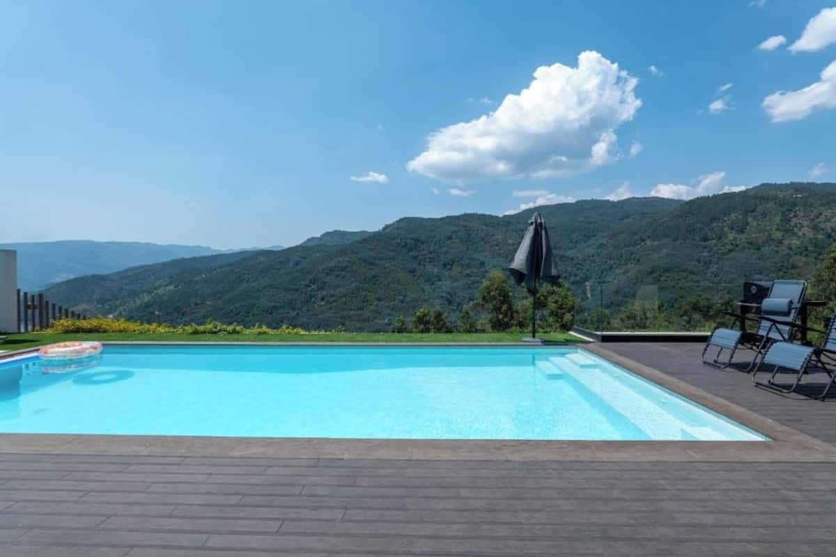 A swimming pool with a view of mountains, surrounded by lounge chairs and an umbrella.