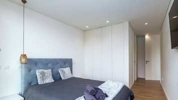 A modern bedroom with a large bed, a grey upholstered headboard, and a flat-screen TV mounted on the wall.
