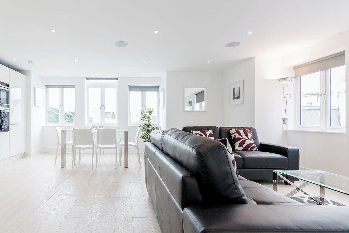 A modern living room with a leather sofa, a glass coffee table, and a dining area with white chairs and a glass table.