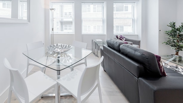 A modern living room with a glass dining table, white chairs, a grey sofa, and large windows with blinds. - Walpole Court (London)