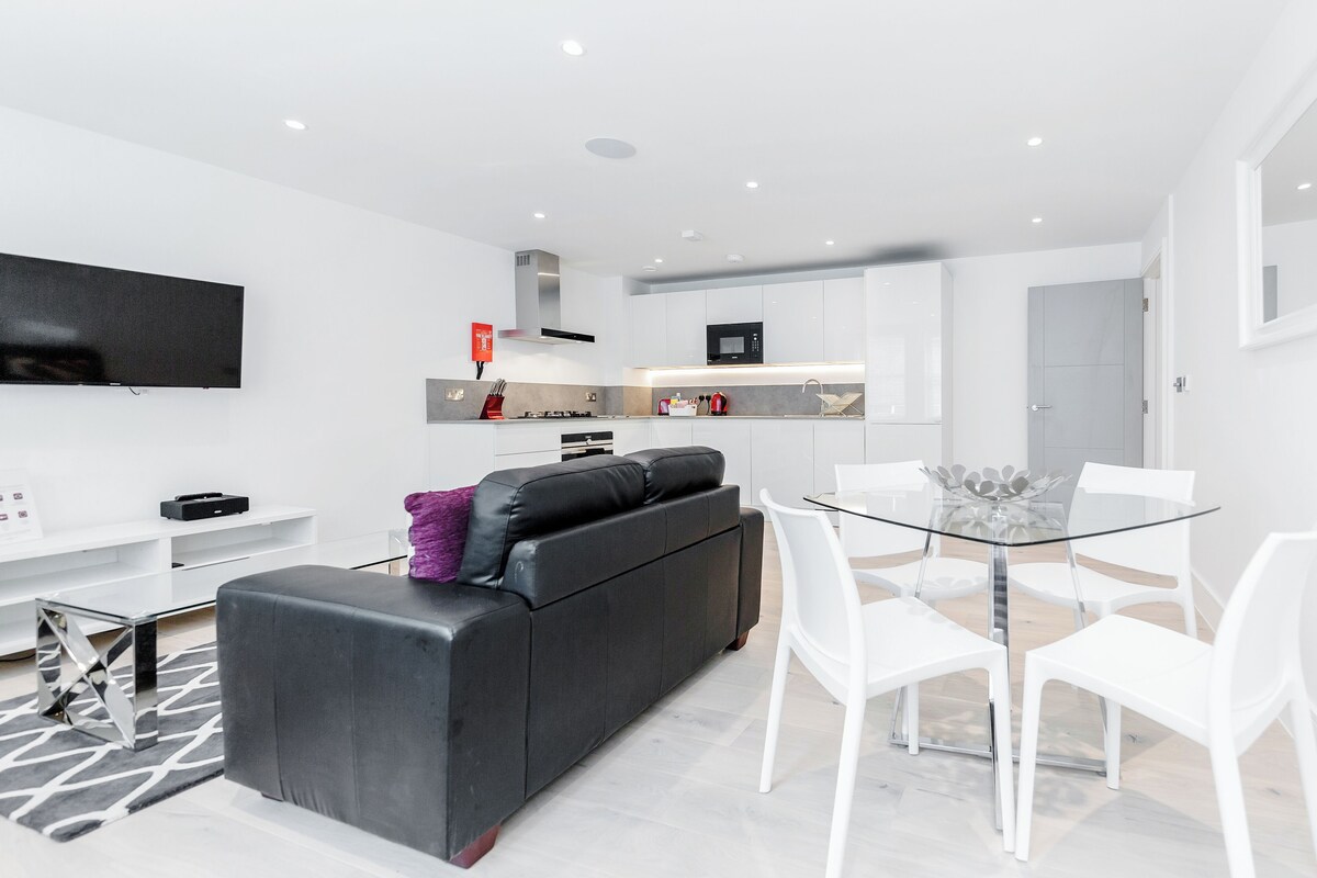 A modern living room with a black sofa, white chairs, a glass dining table, and a kitchen area in the background.