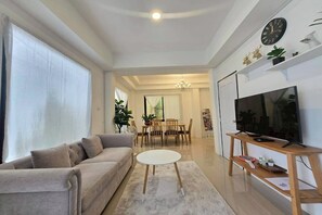 A modern living room with a sofa, a round coffee table, a TV on a wooden stand, and a dining area in the background. - Breezy Bliss Home (Chiang Mai)