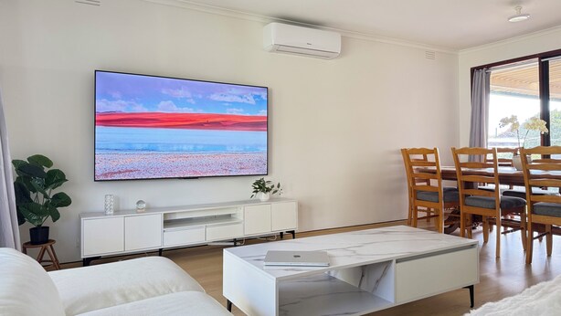 A modern living room with a flat-screen TV, white furniture, a dining area with wooden chairs, and a large window with curtains. - 5 BR House - Private Home on Shared Block (Frankston)