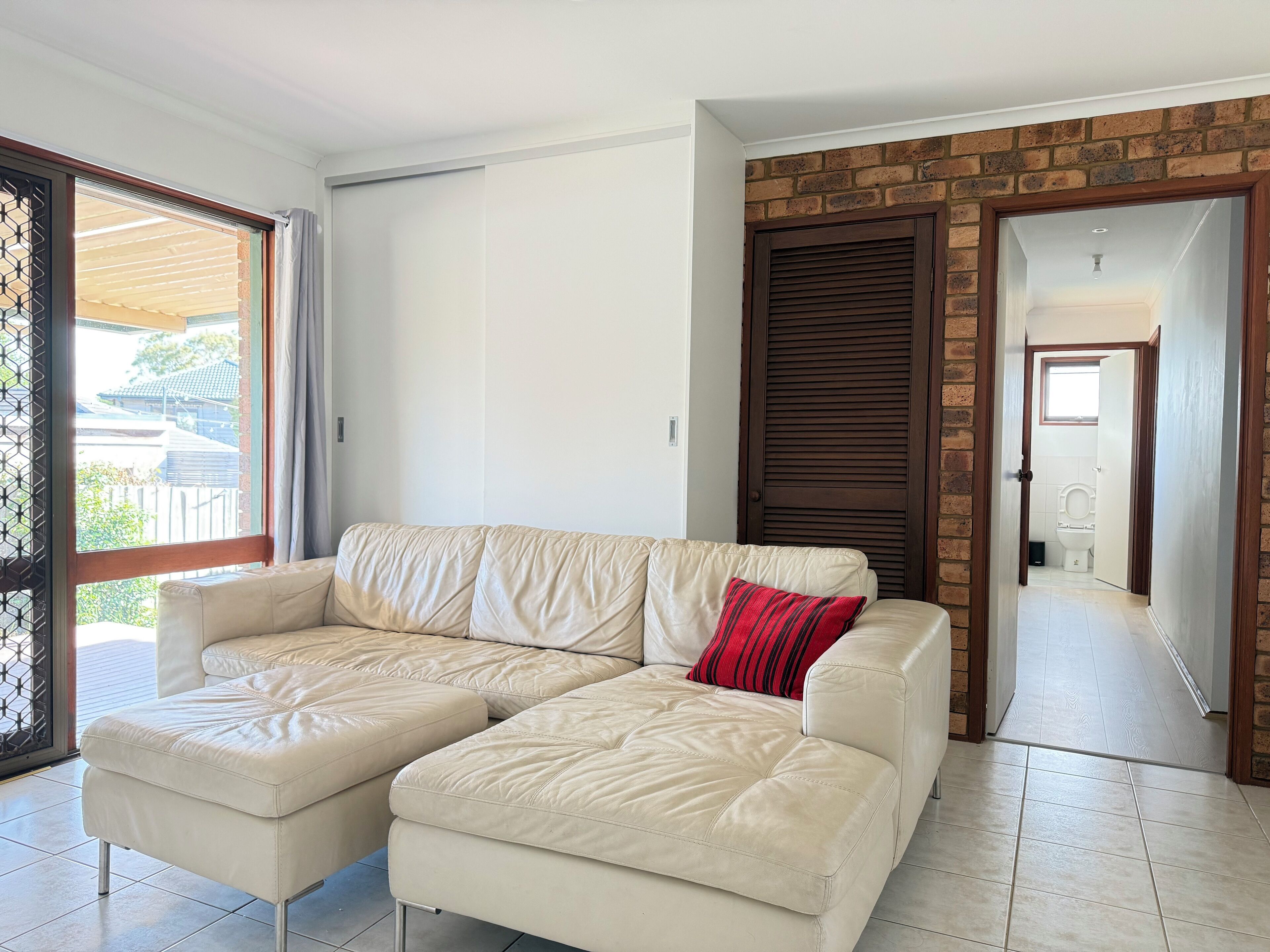 A modern living room with a beige leather sectional sofa, a red pillow, and a view through a glass door to the outside.
