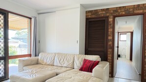 A modern living room with a beige leather sectional sofa, a red pillow, and a view through a glass door to the outside. - 5 BR House - Private Home on Shared Block (Frankston)