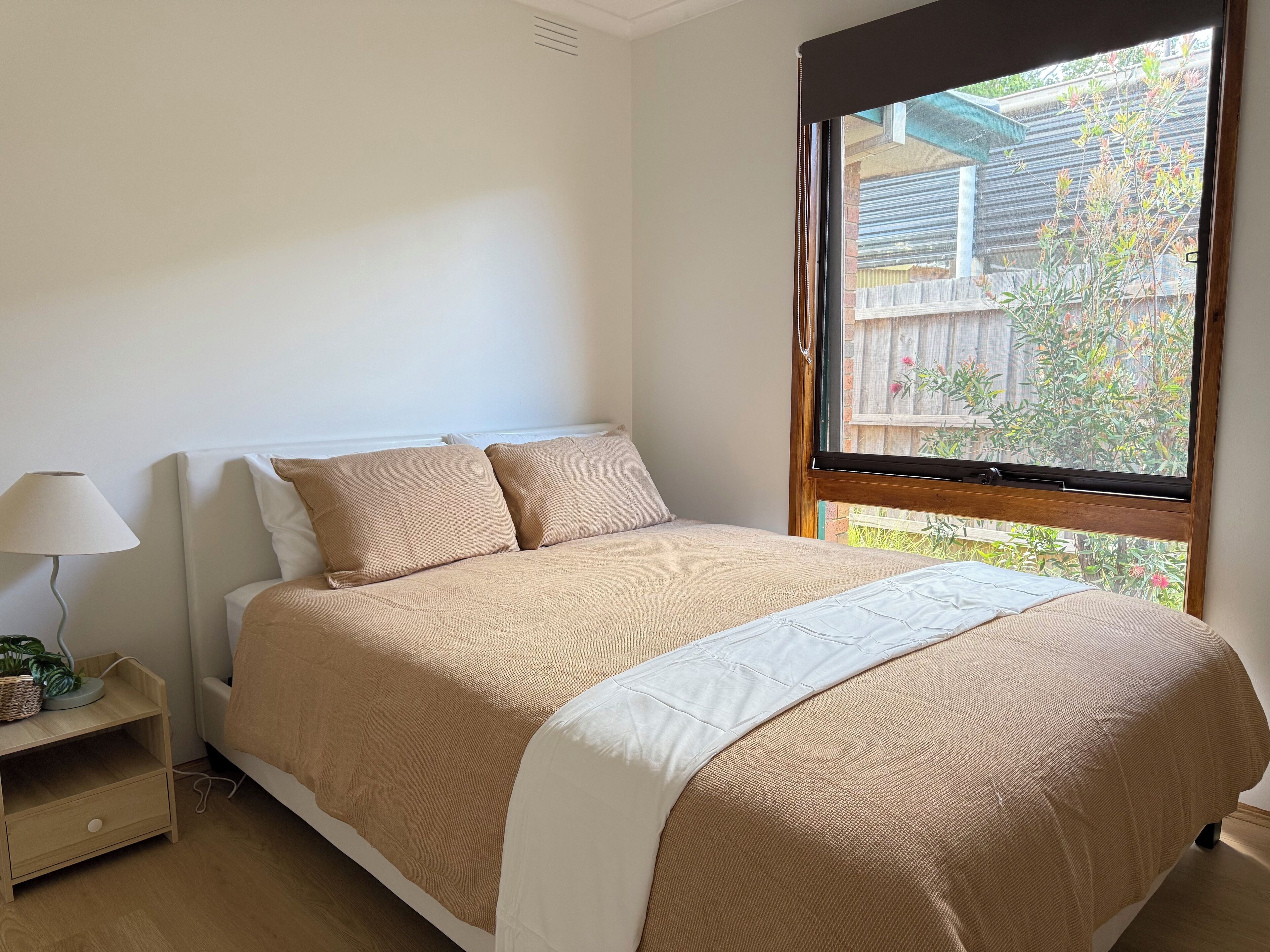 Ein Schlafzimmer mit Bett, Nachttisch, Lampe und Fenster mit Blick ins Grüne.