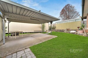 A covered patio with a concrete floor, a small table, and a well-maintained lawn. - 5 BR House - Private Home on Shared Block (Frankston)